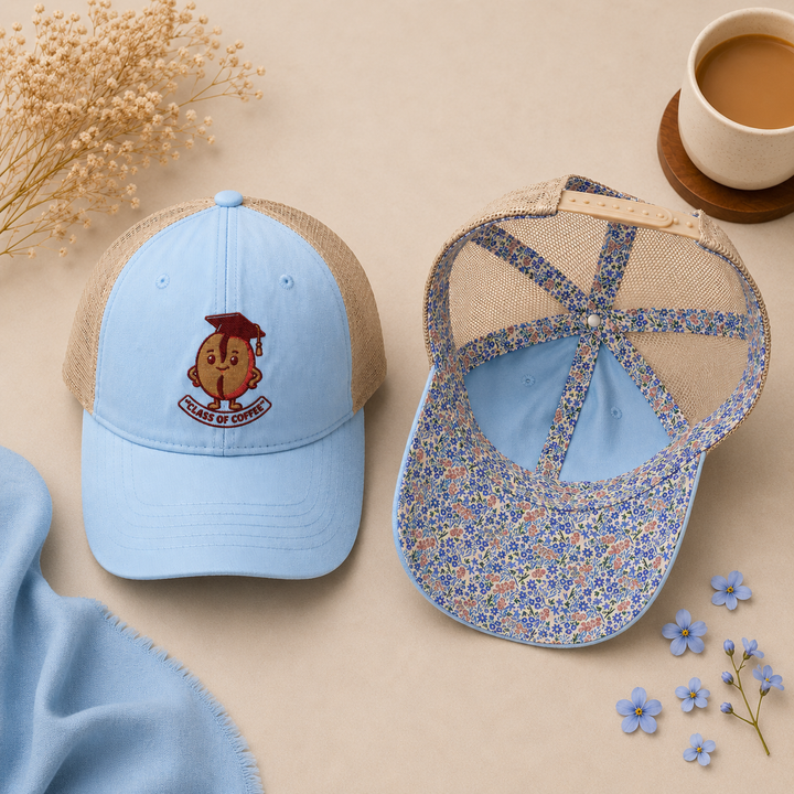 Two blue baseball caps with floral patterns on a beige surface with flowers and a cup of coffee.