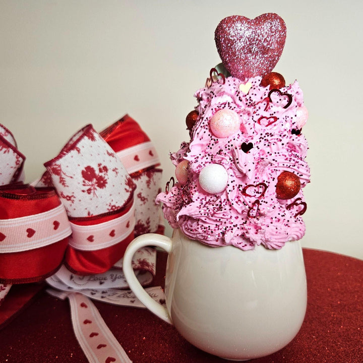 Decorative pink cupcake on a white mug with heart-themed decorations, surrounded by red and white gift bags.