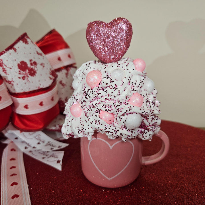 Decorative cup with heart-shaped top and pink ribbon on a red surface