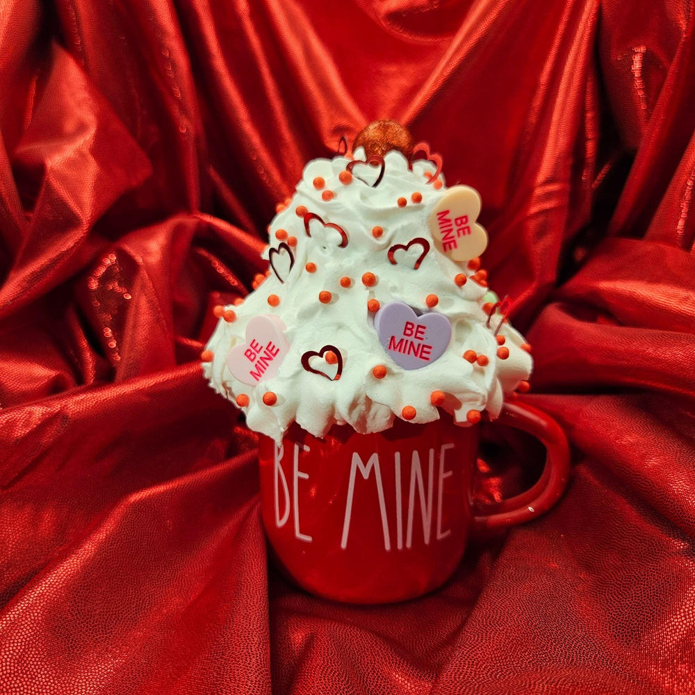 Red mug with 'Be Mine' text, topped with whipped cream and heart-shaped candies on a red fabric background.